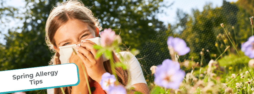 Young girl sitting in a field of wildflowers, sneezing into a tissue, with text overlay that reads “Managing Your Child’s Asthma: Spring Allergy Tips”. Bright green trees and flowers in the background suggest a sunny spring day.